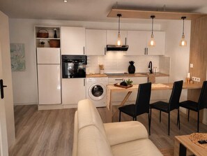 A compact kitchen with a washing machine, a dining area with a table and chairs, and a sofa. - Appartement avec terrasse privée , parking privé, à 7 min des Thermes de Saujon (Saint-Romain-de-Benet)