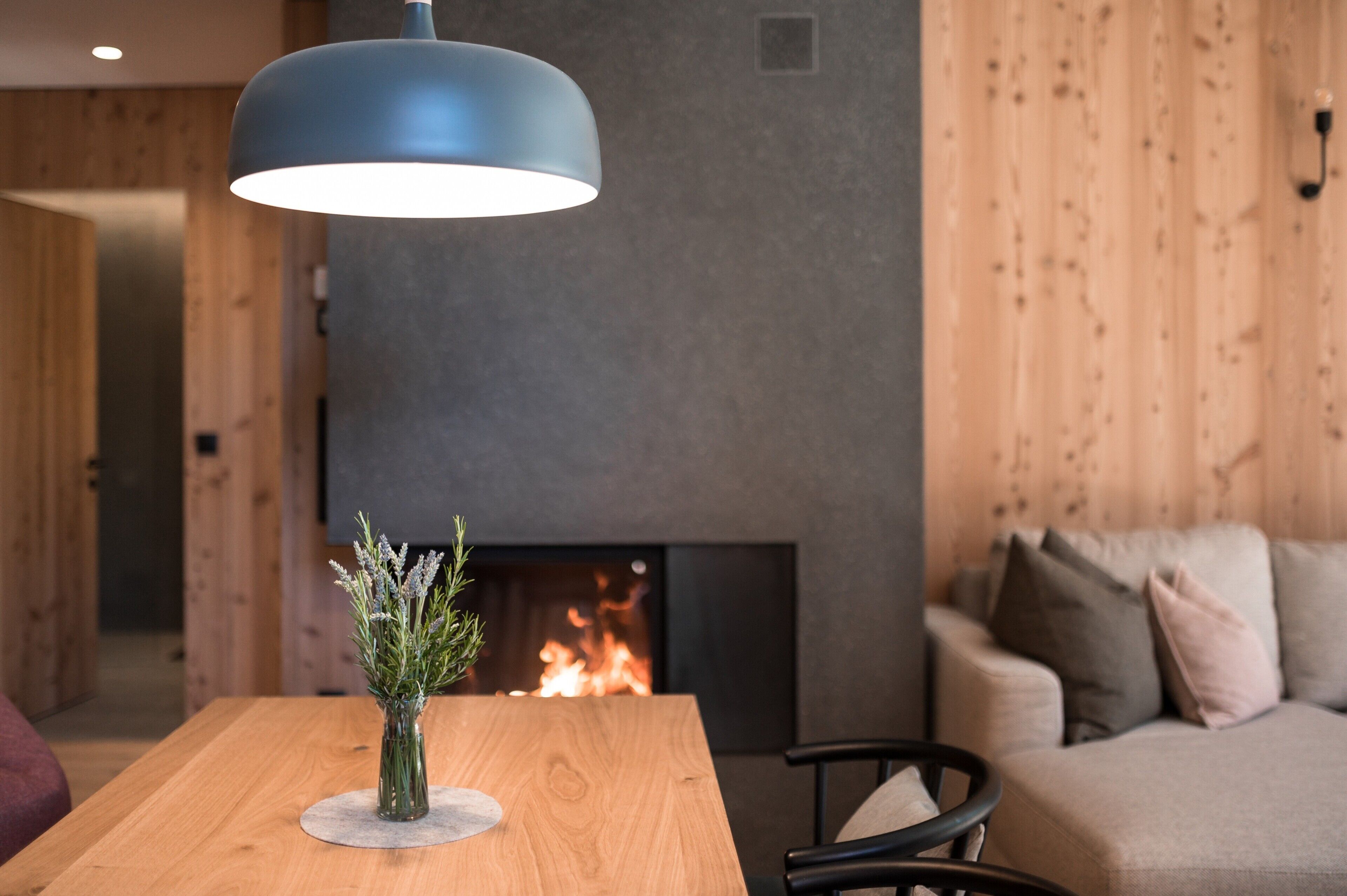 A modern living room with a wooden table, a blue pendant light, a fireplace, and a sofa.