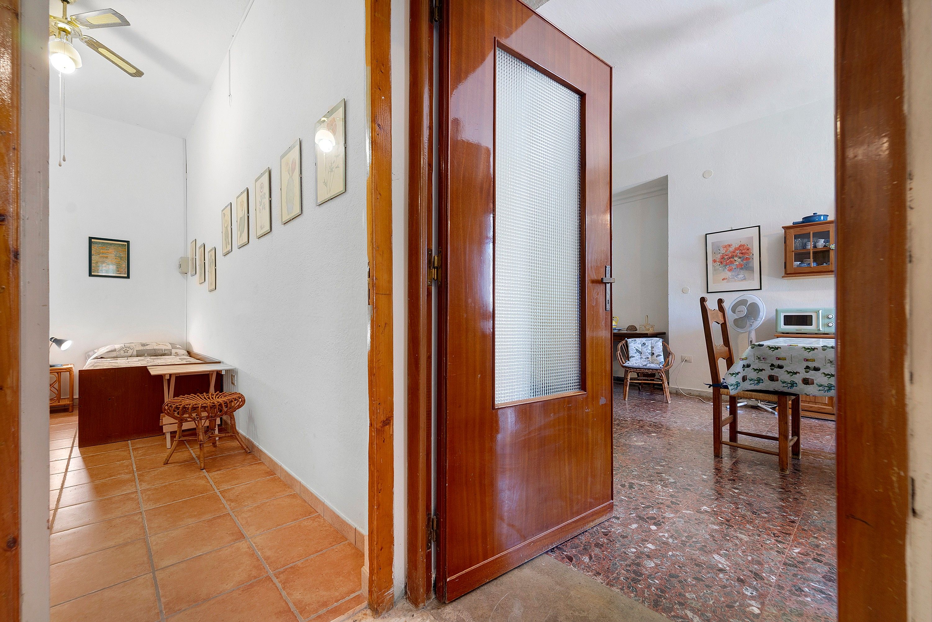 Un couloir avec une porte en bois, un sol carrelé, un lit, une chaise et une table sur laquelle se trouve un ventilateur.