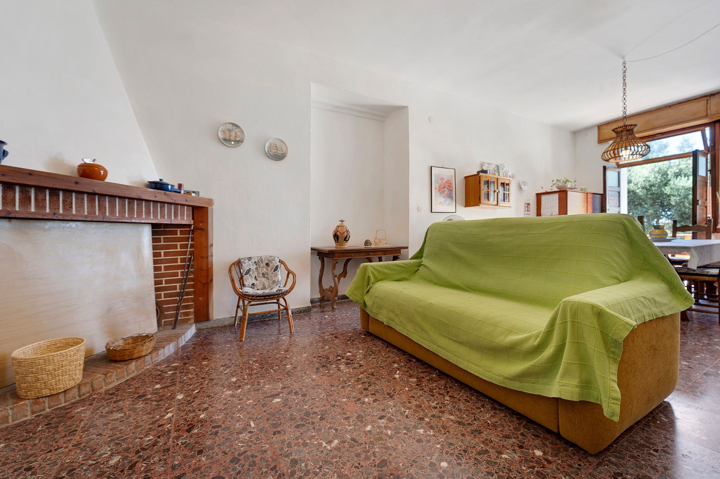 Ein Wohnzimmer mit Kamin, einer mit Grün bezogenen Couch, einem Korbstuhl und einem Essbereich mit Holztisch und Stühlen.