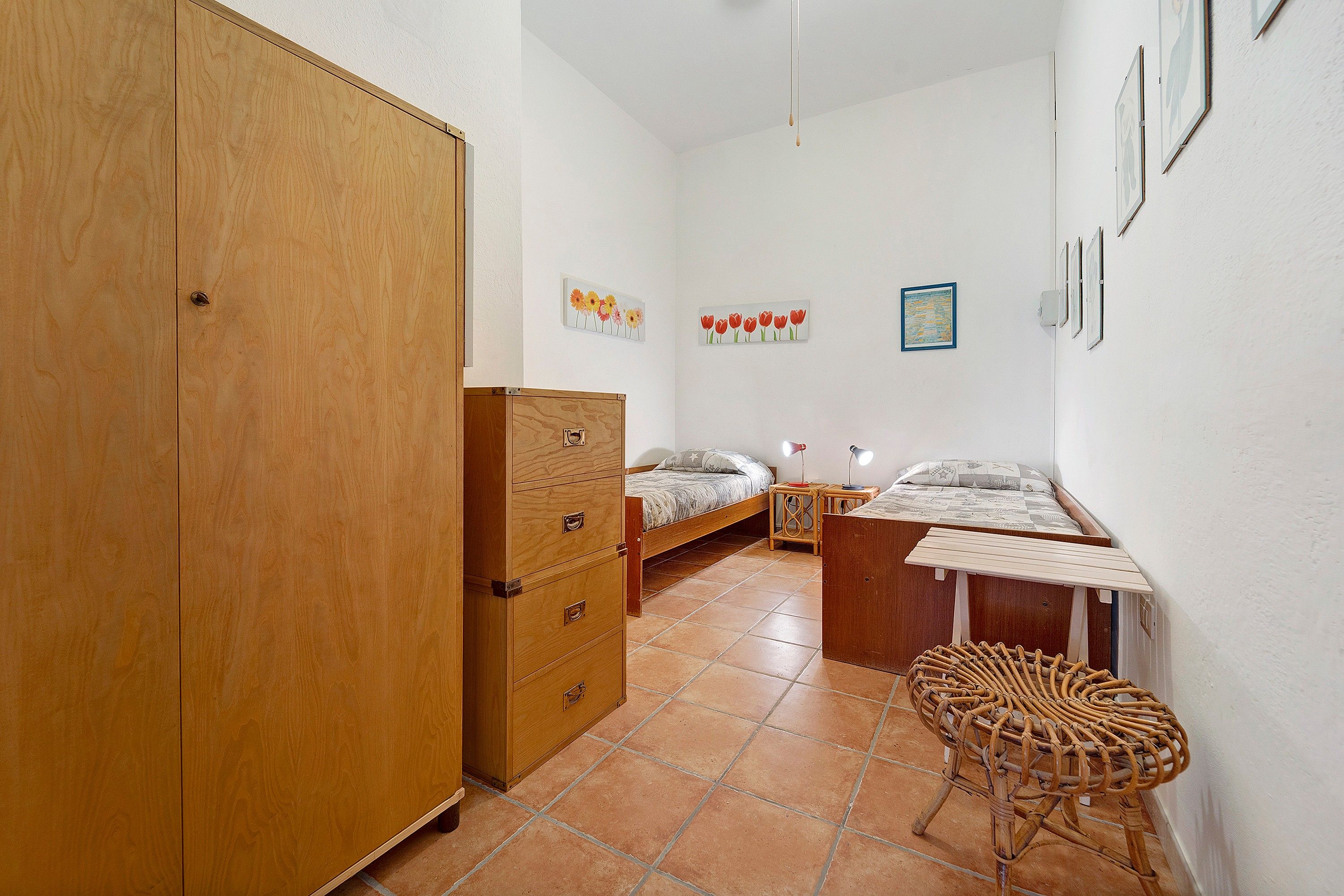 Ein Schlafzimmer mit einem hölzernen Kleiderschrank, einem Bett mit weißem Kopfteil, einem kleinen Tisch mit Lampe und einem Korbstuhl.