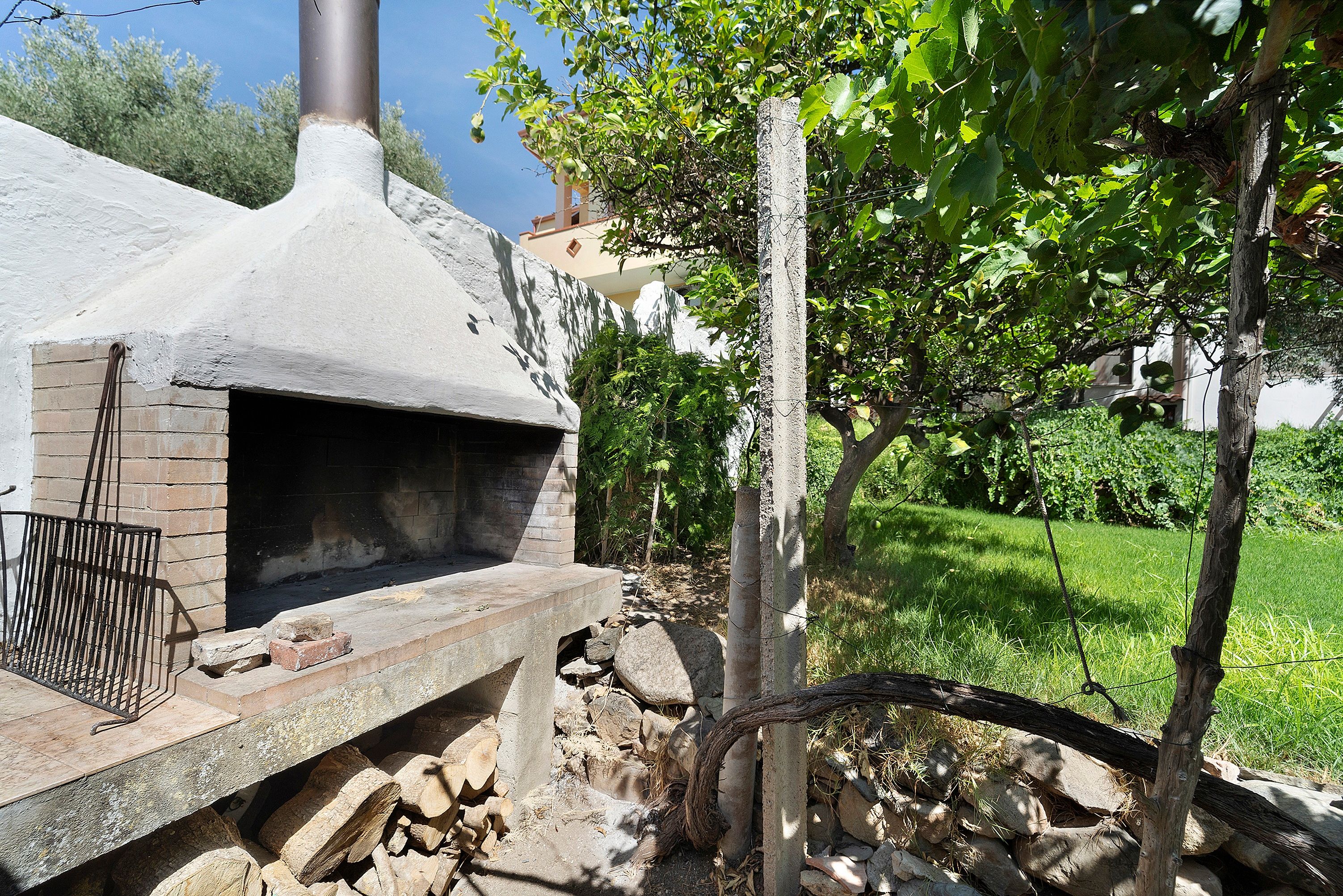 Un four extérieur en briques, doté d’une cheminée, entouré de verdure et bordé d’un mur de pierres.