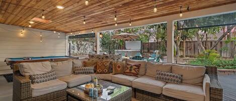 An outdoor living area with a pool table, wicker furniture, and a ceiling with exposed beams and hanging lights.