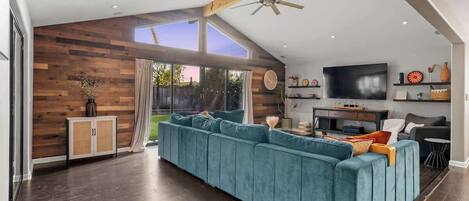 Una sala de estar moderna con un amplio sofá seccional azul, paneles de madera, un ventilador de techo y un televisor de pantalla plana.
