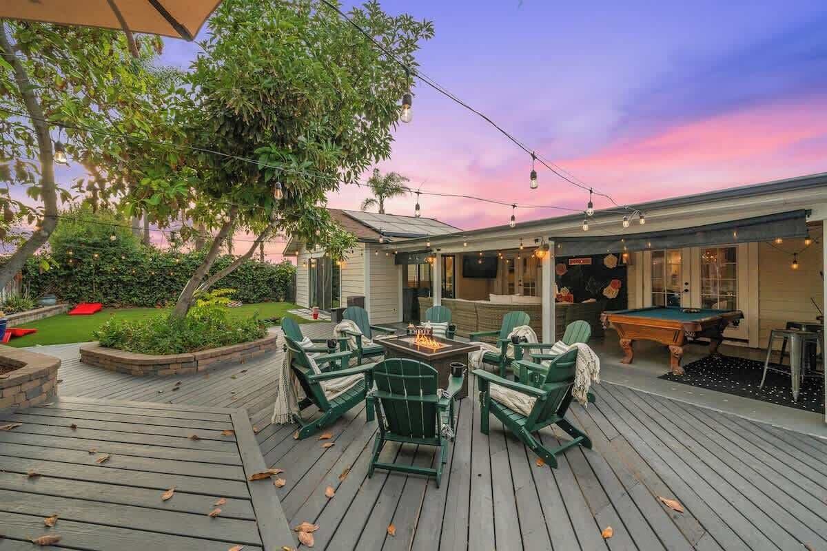 An outdoor deck with green chairs and a fire pit, surrounded by a garden and a building with a pool table.