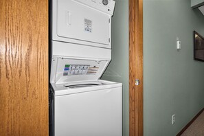 Eine weiße Waschmaschine und ein Trockner stehen übereinander in einer Diele mit einem hölzernen Türrahmen und einer hellgrünen Wand.