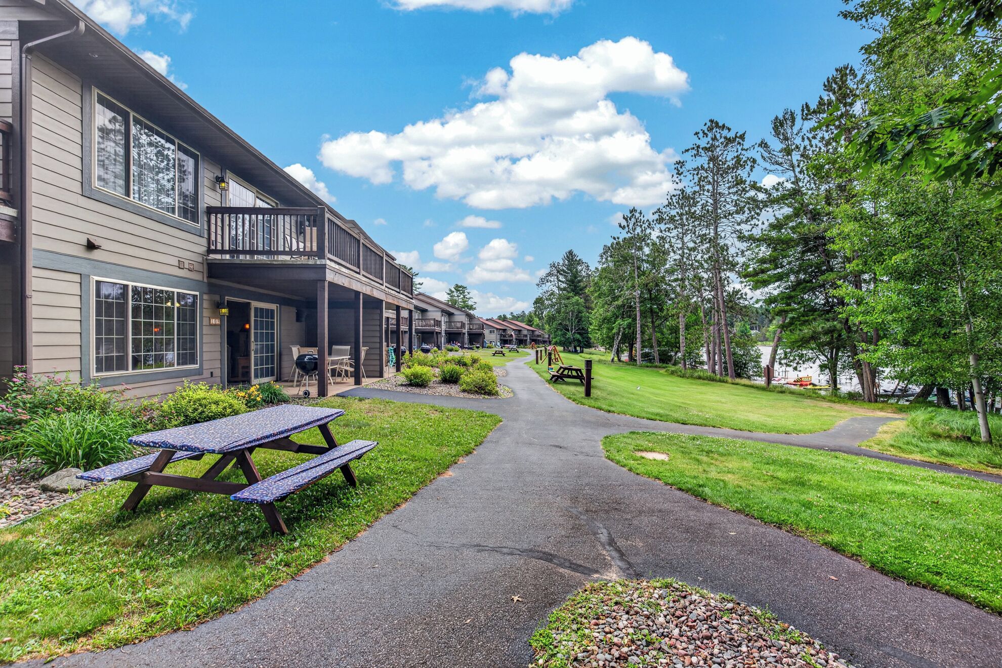 A building with a covered patio, picnic tables, and a pathway leading to a grassy area with trees.