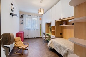 A compact bedroom with a bed, a desk, a chair, a fan, and a window with curtains.