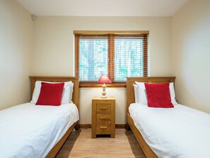 Ein Zimmer mit zwei Betten, jeweils mit einem hölzernen Kopfstück, weißer Bettwäsche und roten Kissen. Dazwischen steht ein kleiner Nachttisch mit einer Lampe.