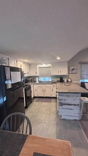 A modern kitchen with white cabinets, a black refrigerator, and a dining area with a table and chairs. - 1495 Ocean Lakes Golf Cart ⛱️🏝️🩱🌊👙☀️🐚 (Myrtle Beach)