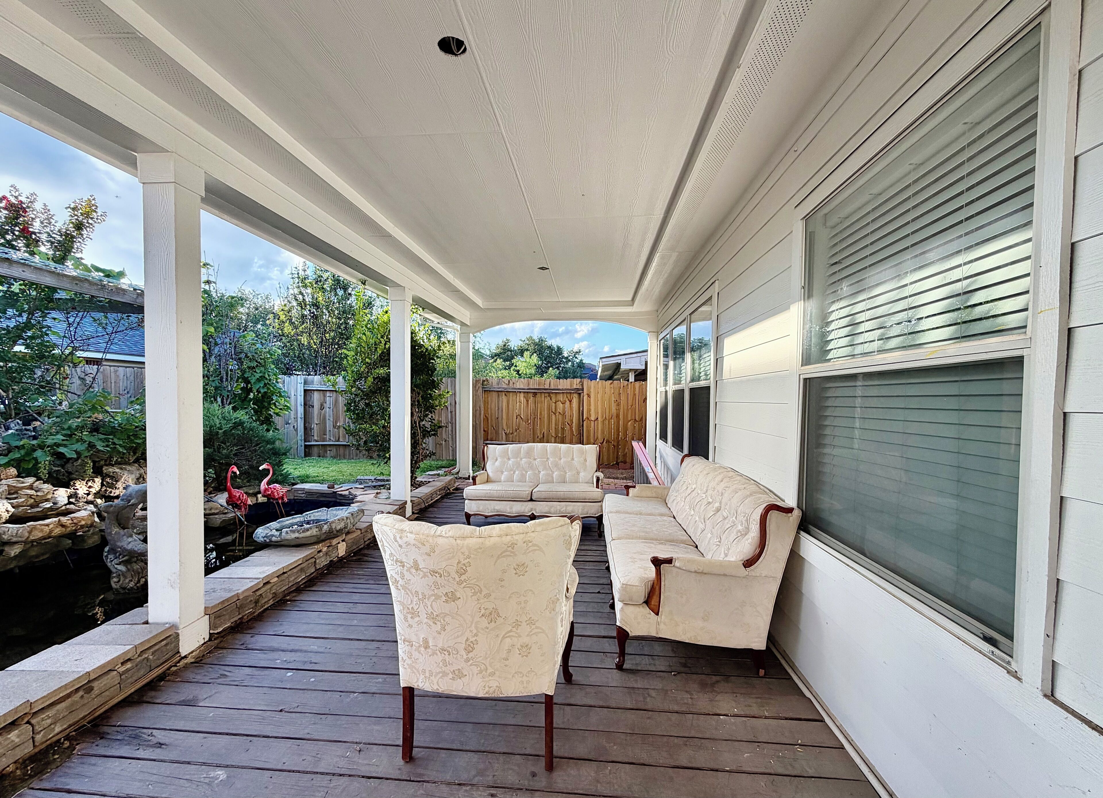 A covered porch with a wooden deck, two white cushioned chairs, and a small pond with flamingos.