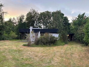 A modern house with a black roof, surrounded by a grassy yard and dense trees. - Bright 1960s house with natural plot near beach (Højbyb)