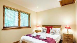 A neatly made bed with white linens and a purple throw blanket, flanked by two bedside tables with lamps, in a room with wooden flooring and a large window with blinds. - Cosy apartment for 4 people with TV, patio and pets allowed (Coldstone)