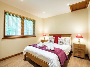 Ein ordentlich bezogenes Bett mit weißen Leinen und einer violetten Tagesdecke, flankiert von zwei Nachttischen mit Lampen, in einem Zimmer mit Holzboden und einem großen Fenster mit Jalousien.
