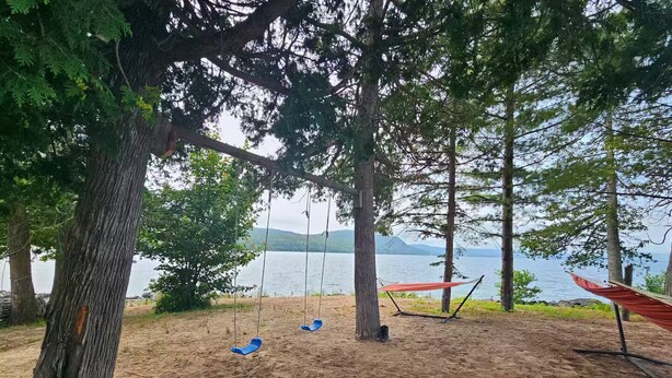 A sandy beach with a hammock and a swing set, surrounded by trees and a lake.