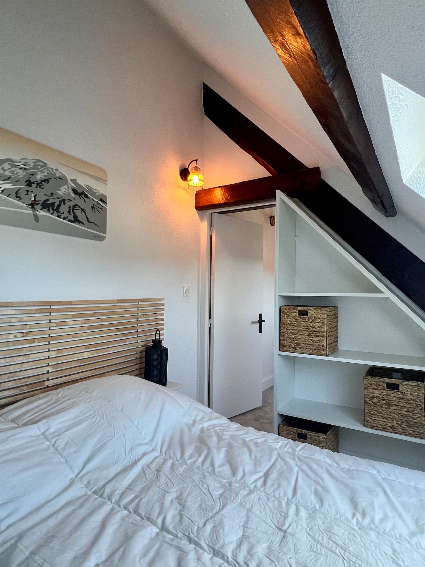 Een slaapkamer met een bed, wandlamp, houten hoofdbord en een ingebouwde wandplank met manden.