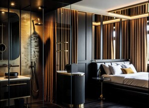 Un dormitorio moderno con cabina de ducha de vidrio, una mesita de noche redonda, una cama con múltiples almohadas y una mesita auxiliar.