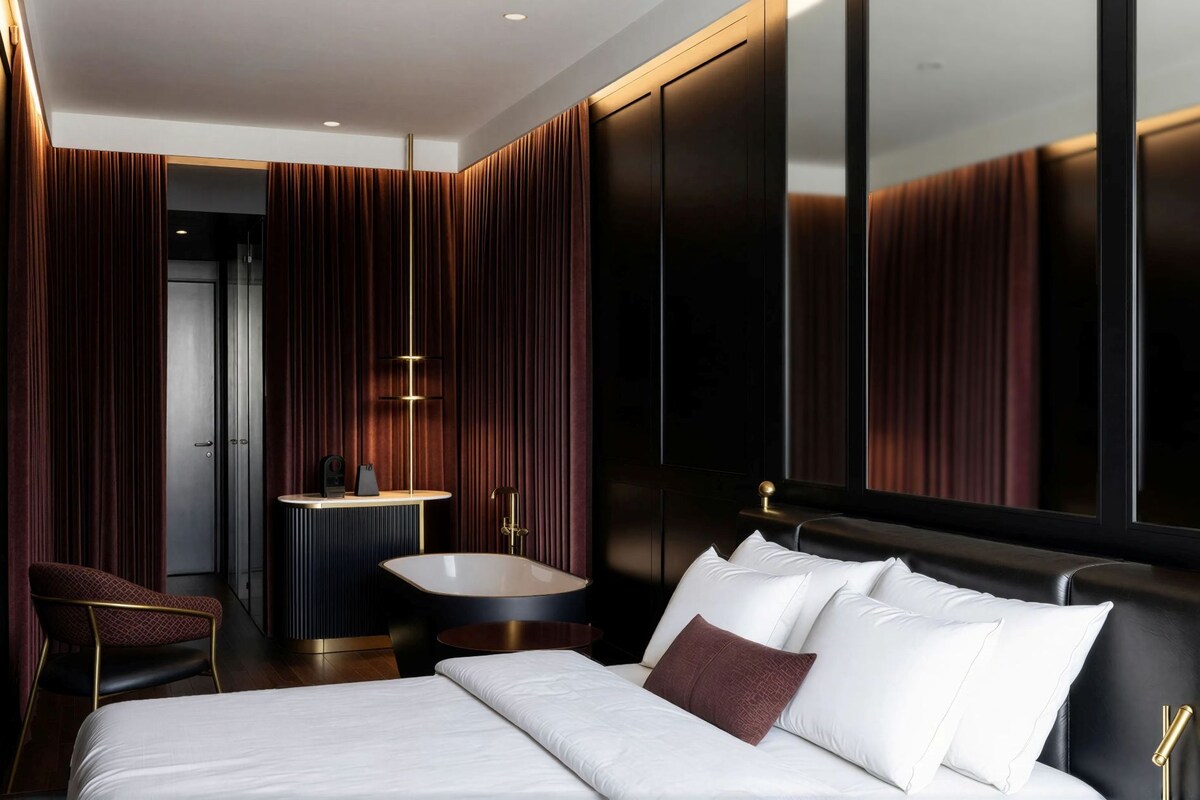 A modern hotel room with a large bed, a sitting area with a chair, a vanity with a mirror, and a bathtub.