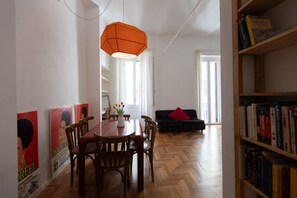 Ein Essbereich mit einem Holztisch und Stühlen, einer hängenden orangefarbenen Leuchte, einem Bücherregal und einem Sofa mit einem roten Kissen.