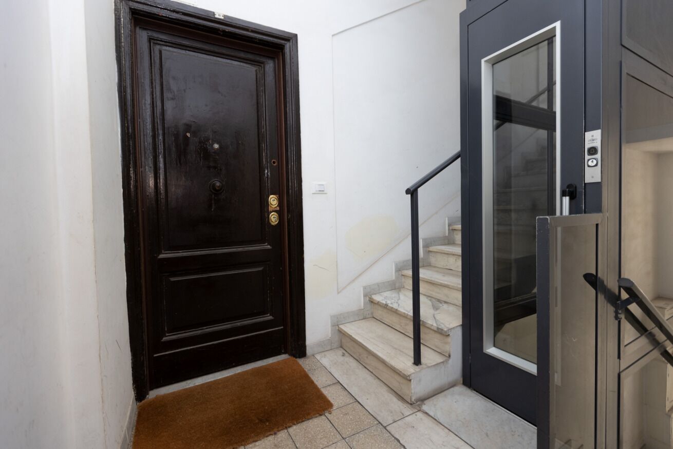 A black door with a brass handle, a brown doormat, and a staircase with a glass door leading to an elevator.