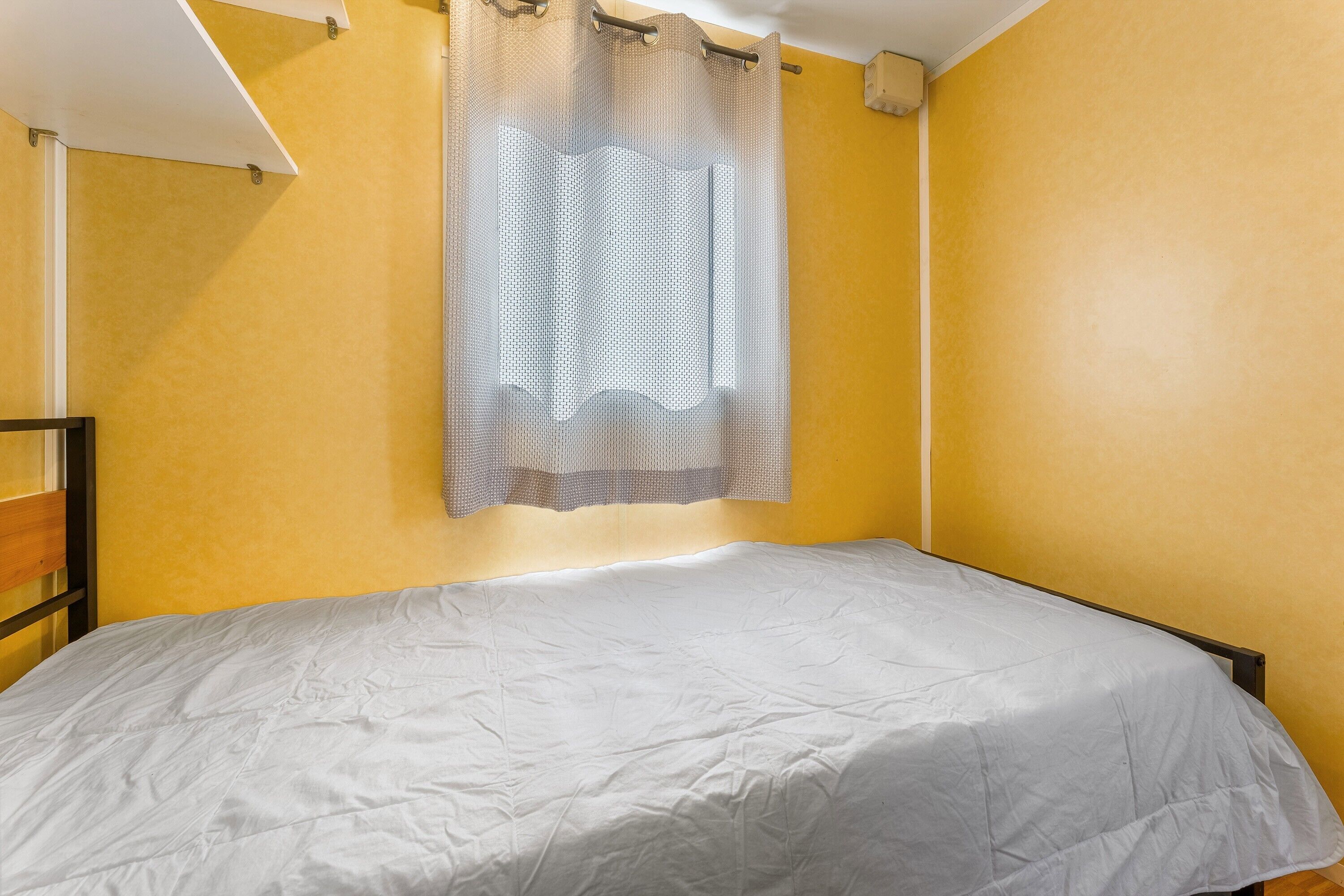 A single bed in a room with yellow walls, a white ceiling, and a window with a sheer curtain.