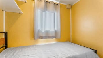 A single bed in a room with yellow walls, a white ceiling, and a window with a sheer curtain.