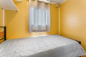 A single bed in a room with yellow walls, a white ceiling, and a window with a sheer curtain.