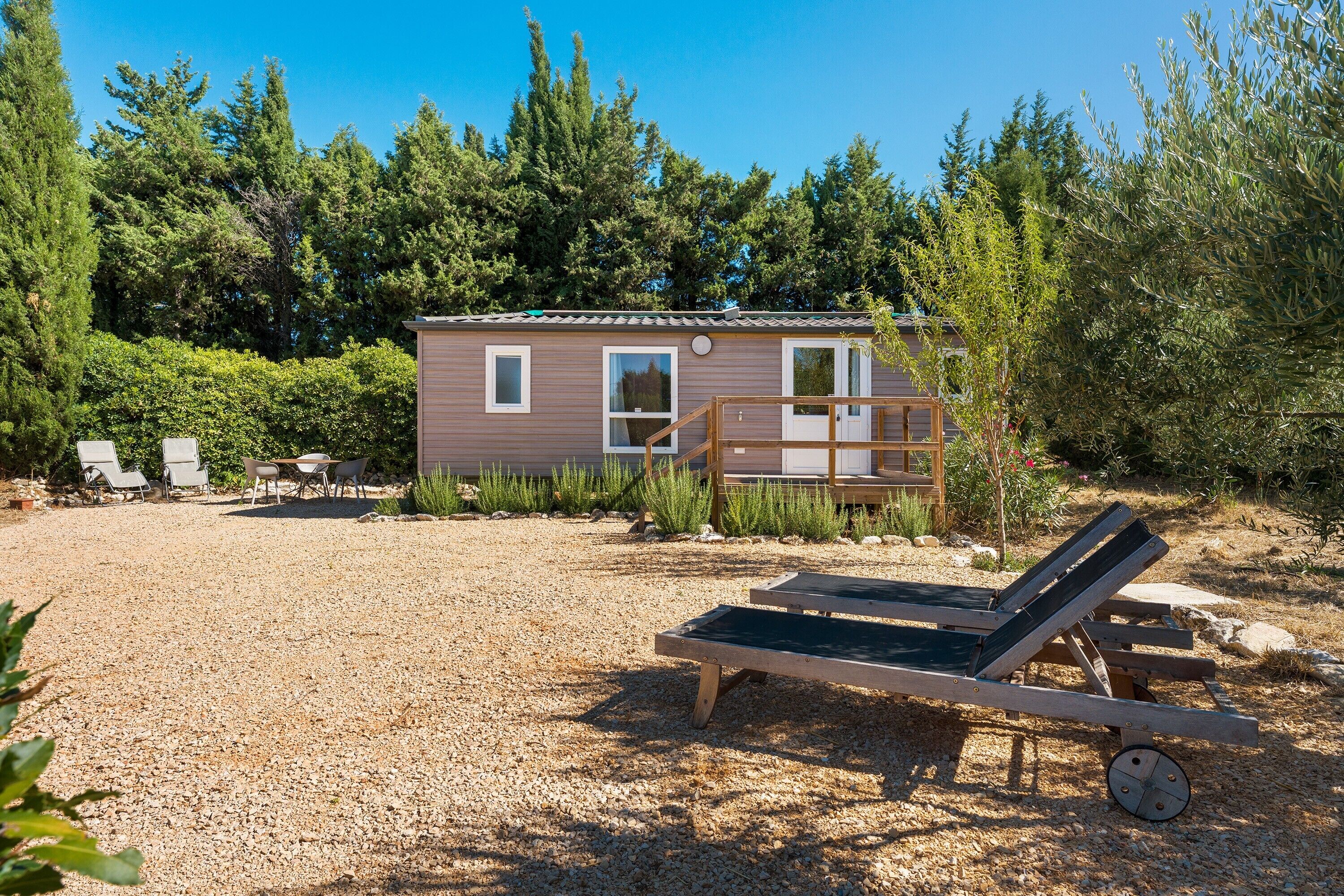 Une petite cabane dotée d’une terrasse, entourée de gravier et de verdure.