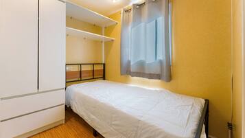 Una habitación pequeña con una cama individual, piso de madera, colcha blanca y pared amarilla.