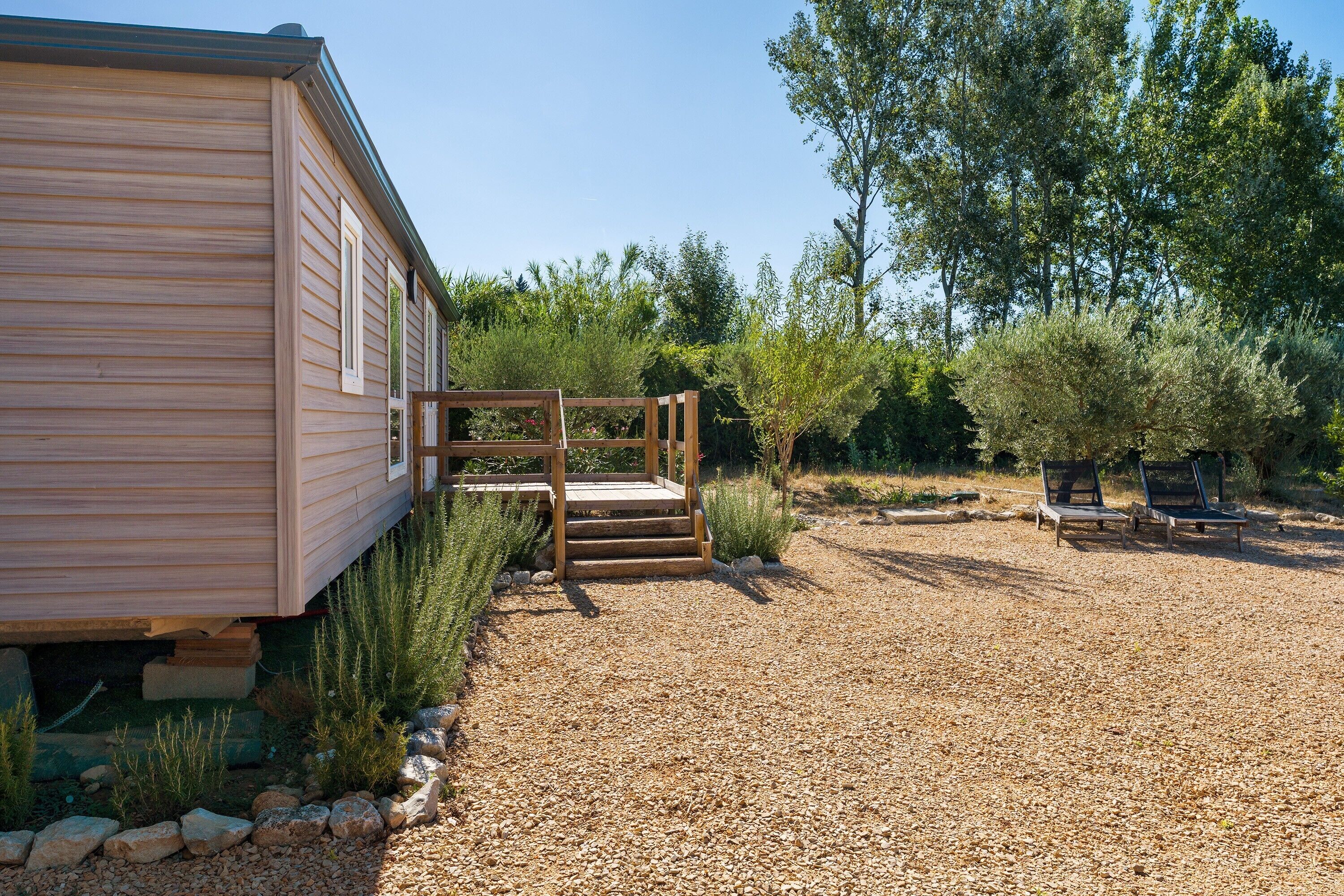 A wooden cabin with a gravel driveway, wooden steps leading to a deck, and a garden with various plants and a bench.