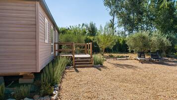 A wooden cabin with a gravel driveway, wooden steps leading to a deck, and a garden with various plants and a bench.