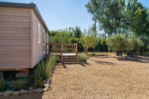A wooden cabin with a gravel driveway, wooden steps leading to a deck, and a garden with various plants and a bench.