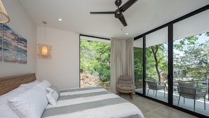 Una camera da letto con un letto, un ventilatore a soffitto, un'ampia finestra con tende e un balcone con sedie.