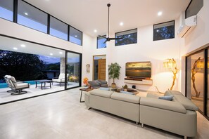 A modern interior with a large sofa, a dining table, and a TV. There is a view of the outdoors through large windows. - New Listing • Casa Azul Oasis • Pacifico 4BR Villa • Private Pool (Playas del Coco)