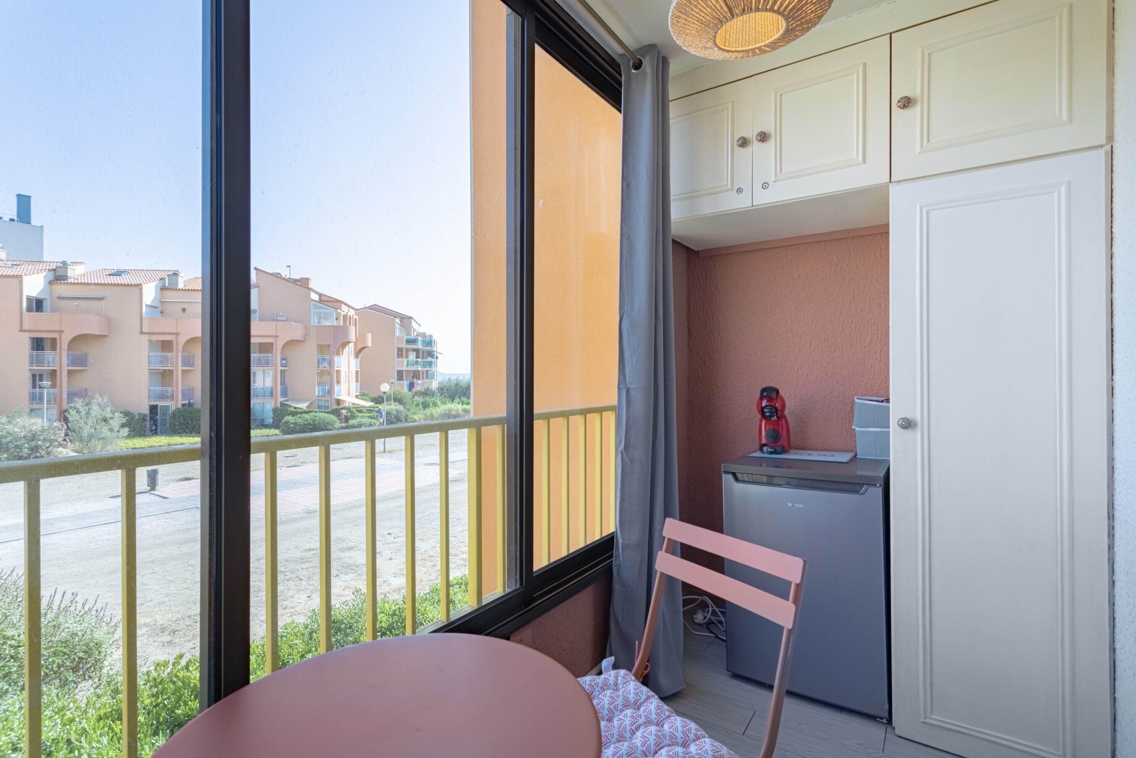 A balcony with a view of a residential area, a small kitchen area with a refrigerator, and a chair.