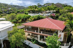 A residential building with a red roof, surrounded by lush greenery and other houses in the background. - New Listing • 2BR Ocean View Condo • Coco Joya • Walk to the Beach (Playas del Coco)