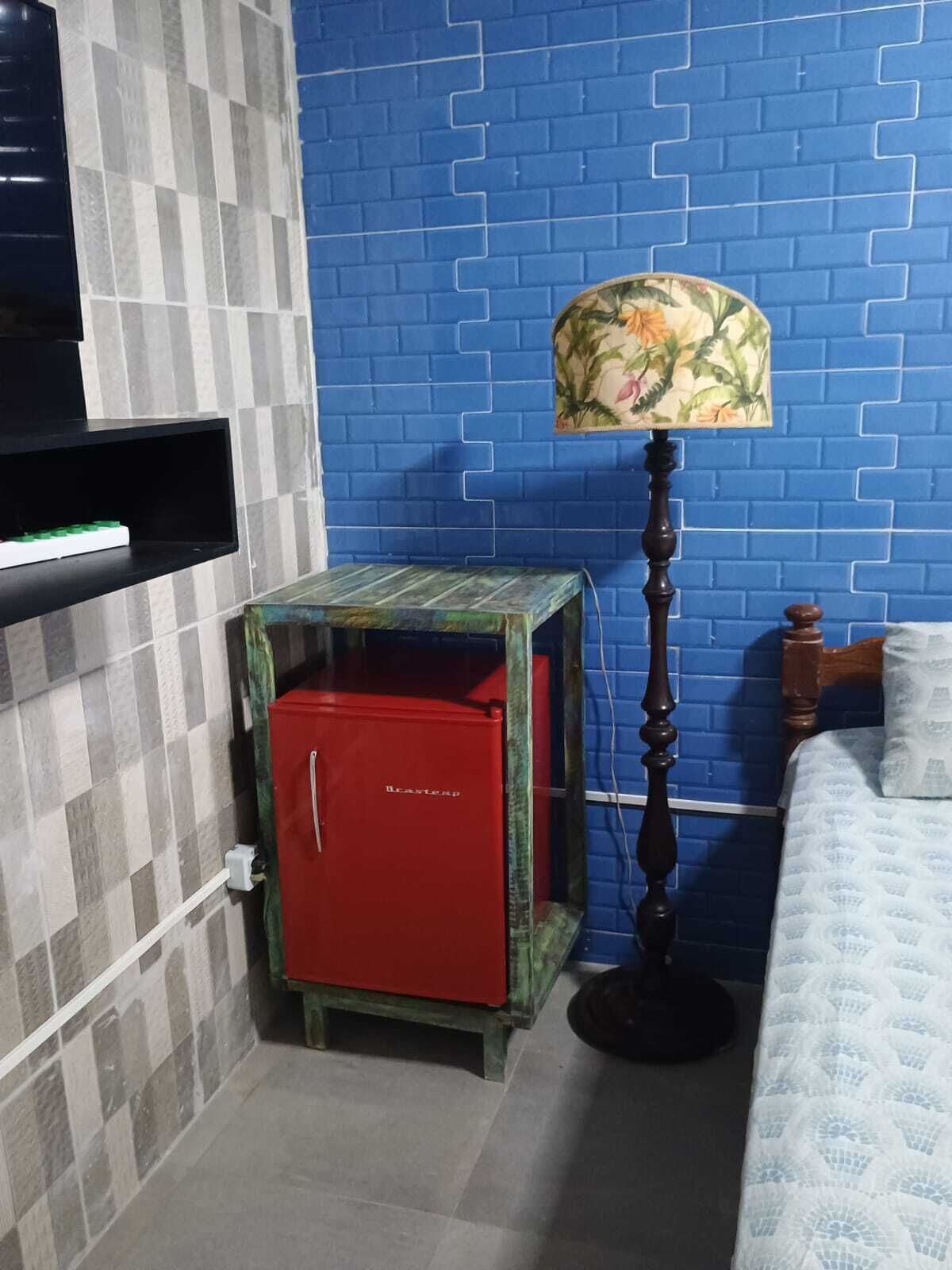 Una habitación con pared de ladrillo azul, una cama con ropa blanca, un armario rojo, una lámpara de pie con pantalla floral y un estante con un televisor.