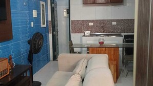 Sala de estar con sofá, ventilador, una mesita y una kitchenette con alacenas y salpicadero de azulejos.