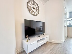 Una sala moderna con un televisor de pantalla plana grande sobre un mueble blanco, un reloj de pared decorativo y un pequeño jarrón con ramas.