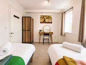 A hotel room with two beds, a wooden wardrobe, a desk with a chair, a painting on the wall, and a window with curtains. - Modern 3BDR Family House in North Stifford (North Stifford)