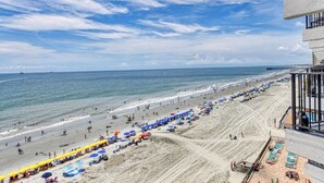 Une plage avec des parasols et des chaises longues, une jetée au loin et un immeuble doté de balcons donnant sur la plage.