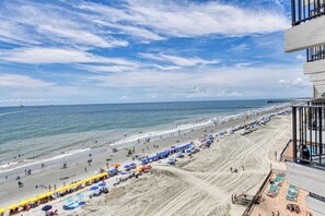 Ein Strand mit Sonnenschirmen und Liegestühlen, ein Pier in der Ferne und ein Gebäude mit Balkonen mit Blick auf den Strand.