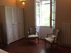 A room with a large wardrobe, two chairs, a radiator, and a window with a view of greenery. - Renovated Provençal farmhouse in a quiet location—2 hectares of wooded land (Sarrians)
