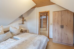 Un dormitorio con techo inclinado, una cama con ropa de cama estampada, una mesita de noche con lámpara y un armario de madera.