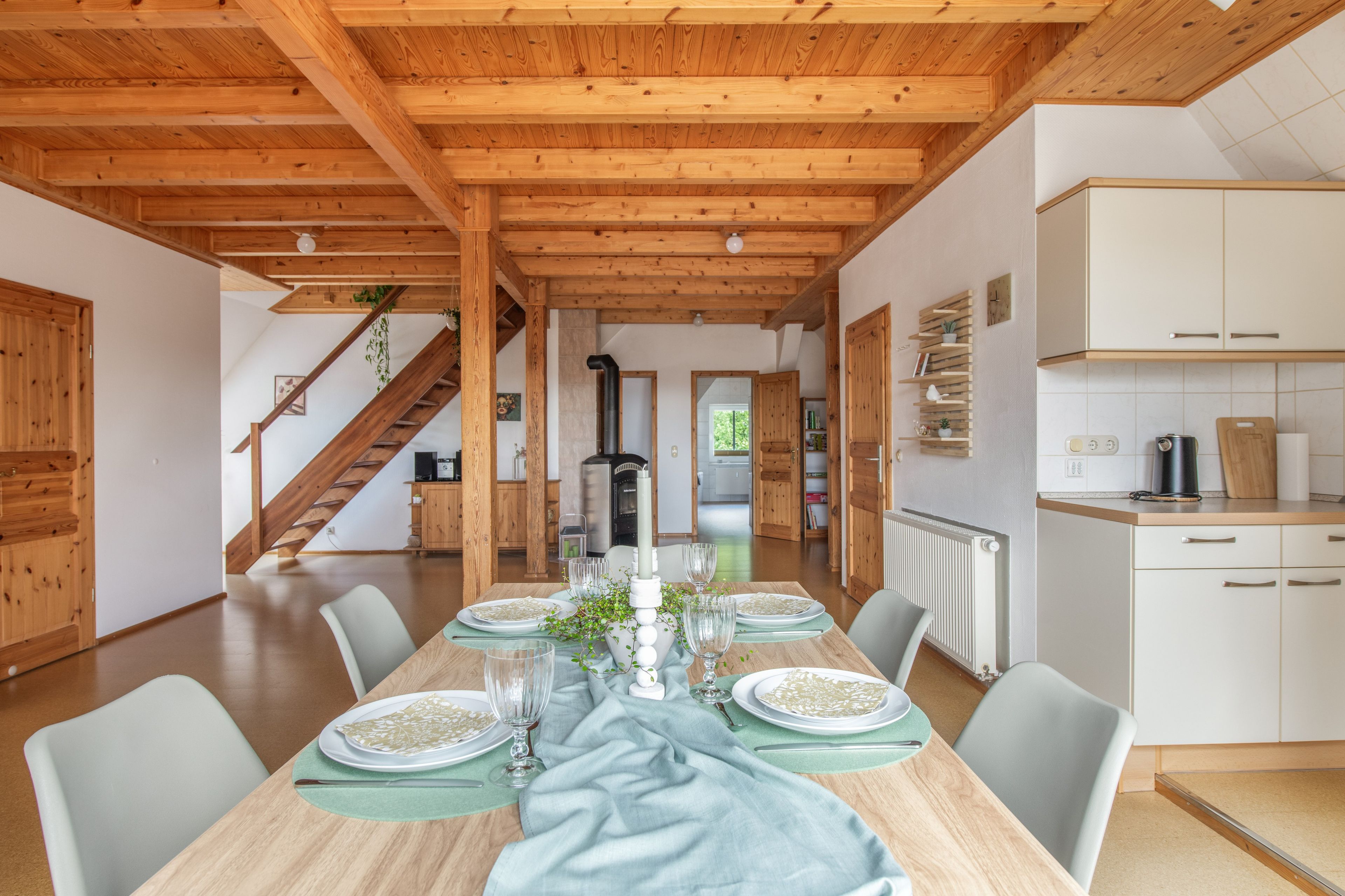 A dining area with a long wooden table set for a meal, flanked by chairs, with a staircase leading to another room.