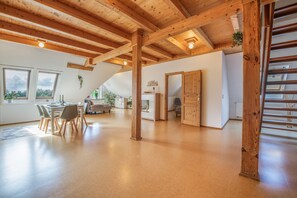 A spacious room with a high ceiling, wooden beams, a dining area with a table and chairs, and a view through an open door.