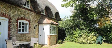 A traditional stone house with a thatched roof, a brick chimney, and a small garden with a lawn and a chair.