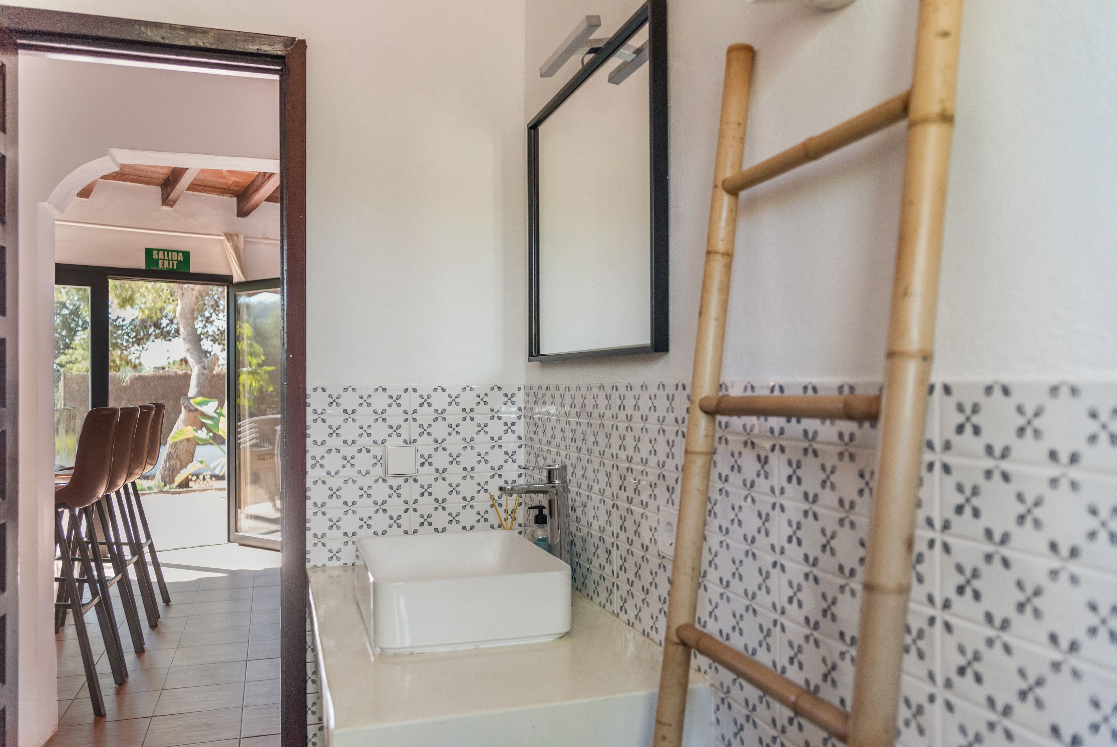A bathroom with a white sink, a mirror, and a bamboo ladder. There is a door leading to an outdoor area with chairs and a view of trees.