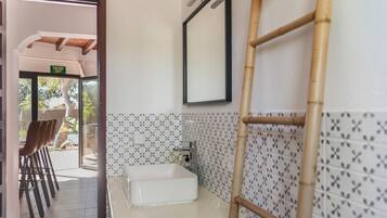 A bathroom with a white sink, a mirror, and a bamboo ladder. There is a door leading to an outdoor area with chairs and a view of trees.