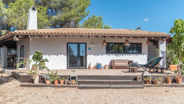 Ein Haus mit Ziegeldach, einer überdachten Terrasse mit Bank und Tisch sowie verschiedenen Kübelpflanzen.